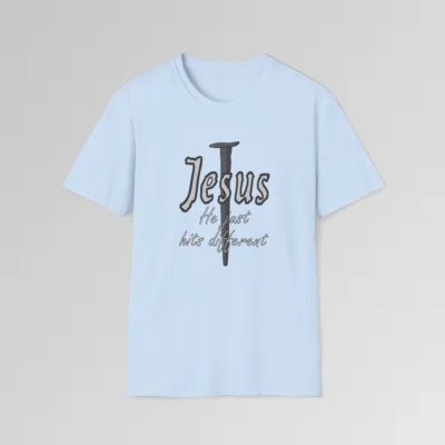 Light blue colored softstyle tee, with a crucifixion spike, and the name "Jesus" in big print over top of it. Beneath Jesus, it says "He just hits different", in a cursive font. The font is silver, with a dark grey border.