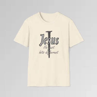 Natural colored softstyle tee, with a crucifixion spike, and the name "Jesus" in big print over top of it. Beneath Jesus, it says "He just hits different", in a cursive font. The font is silver, with a dark grey border.