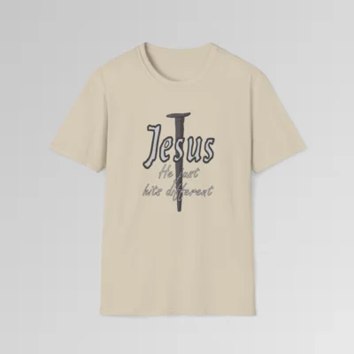 Sand colored softstyle tee, with a crucifixion spike, and the name "Jesus" in big print over top of it. Beneath Jesus, it says "He just hits different", in a cursive font. The font is silver, with a dark grey border.