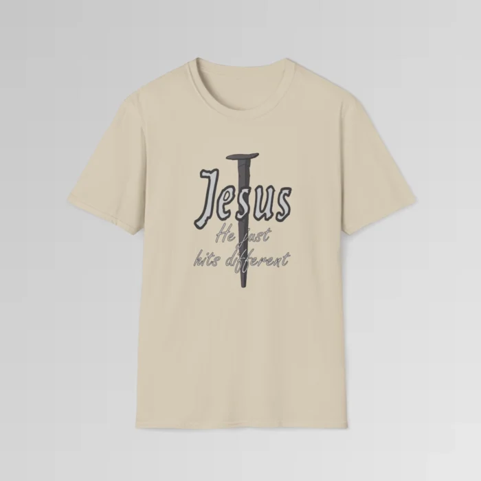 Sand colored softstyle tee, with a crucifixion spike, and the name "Jesus" in big print over top of it. Beneath Jesus, it says "He just hits different", in a cursive font. The font is silver, with a dark grey border.