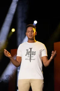 Male model wearing the natural colored Big Baby Gorilla Bear "Jesus Hits Different" Softstyle Tee, with slacks, on a stage with hands lifted out and upward, in a worshipping pose.