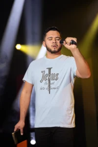 Male model wearing a light blue "Jesus Hits Different" Softstyle Tee, on a stage, holding a mic and Bible.