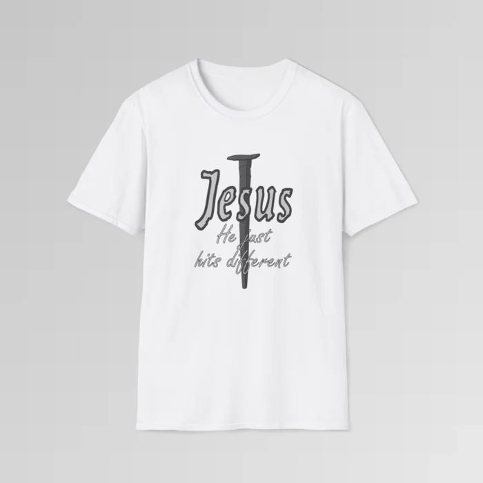 White softstyle tee, with a crucifixion spike, and the name "Jesus" in big print over top of it. Beneath Jesus, it says "He just hits different", in a cursive font. The font is silver, with a dark grey border.