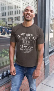 Male model wearing the dark chocolate Big Baby Gorilla Bear "My Wife's Girl's Girl" tee, with jeans, smiling, with a mirrored window background.