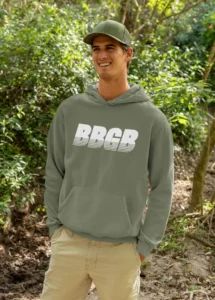 A male model wearing the BBGB Simple Hoodie Dark Colors, Military Green, while standing in nature, smiling, and wearing a green hat and khaki pants.