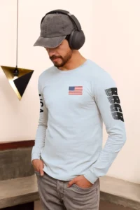 Male Model showing front and left side of the BBGB USA Full Stack Long Sleeve tee, wearing a hat and headphones, in a room. Light Blue.