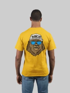 Male model showcasing the back of a gold colored tee, with the Big Baby Gorilla Bear Mascot's face, wearing a white ball cap, and blue and white shades, super imposed on the back of a tee shirt.