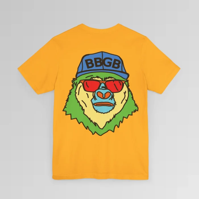 The back of a gold colored shirt, with the Big Baby Gorilla Bear Mascot's face in vibrant colors of light blue, green, yellow, and orange, wearing a blue ball cap, and red shades, super imposed on the back of a t-shirt.