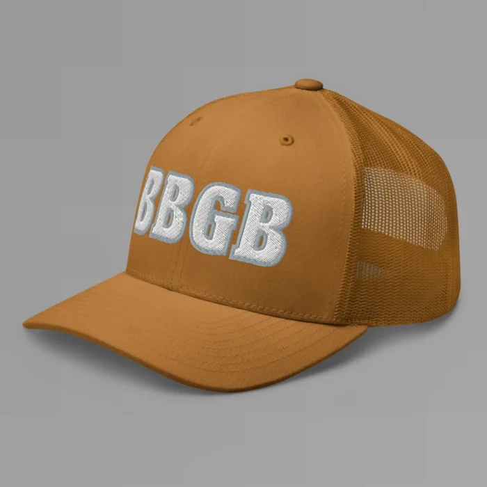 A caramel hat with the letters "BBGB" embroidered on the front in white thread, with a silver border, on a trucker cap with a mesh back. (Left Side View)