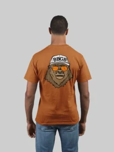 Male model showcasing the back of an autumn colored tee, with the Big Baby Gorilla Bear Mascot's face, wearing a white ball cap, and orange and white shades, super imposed on the back of a t-shirt.