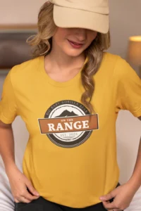 Female model showcasing the front of the "On The Range" t-shirt from Big baby Gorilla Bear, sitting in a room, with a hat on - Gold Color. The graphic is circular, with a mountain range, and a center box with the words "On The Range", and the BGB Company name on top of the circular design, and they year established 2024 on the bottom part of the design.