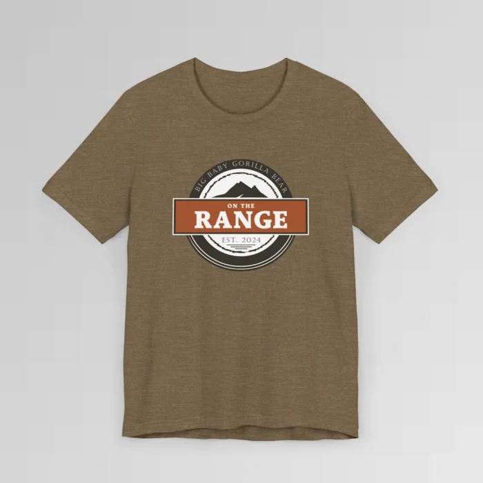 The front of a shirt with a dark brown, and cream white circular design, with the words "Big Baby Gorilla Bear" wrapped on the top part of the circle, and "Est. 2024" on the bottom inner part of the circle. In the center, there is an orange and brown rectangle with the words "On The Range", and a mountain graphic. Color = Heather Olive.