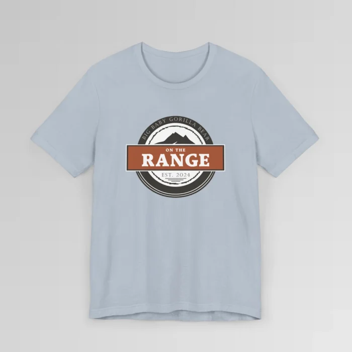 The front of a shirt with a dark brown, and cream white circular design, with the words "Big Baby Gorilla Bear" wrapped on the top part of the circle, and "Est. 2024" on the bottom inner part of the circle. In the center, there is an orange and brown rectangle with the words "On The Range", and a mountain graphic. Color = Light Blue.