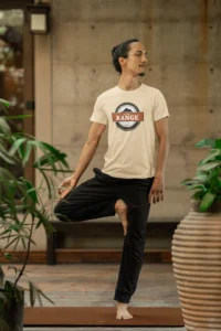Male model showcasing the front of the "On The Range" t-shirt from Big baby Gorilla Bear, in a yoga pose - Soft Cream Color. The graphic is circular, with a mountain range, and a center box with the words "On The Range", and the BGB Company name on top of the circular design, and they year established 2024 on the bottom part of the design.
