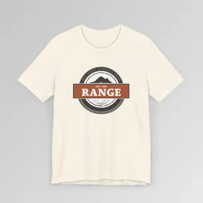 The front of a shirt with a dark brown, and cream white circular design, with the words "Big Baby Gorilla Bear" wrapped on the top part of the circle, and "Est. 2024" on the bottom inner part of the circle. In the center, there is an orange and brown rectangle with the words "On The Range", and a mountain graphic. Color = Natural.