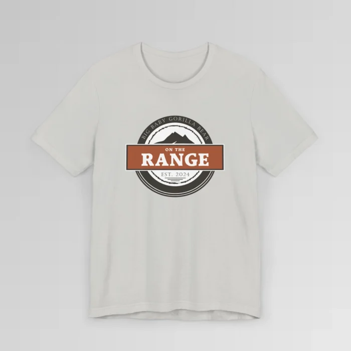 The front of a shirt with a dark brown, and cream white circular design, with the words "Big Baby Gorilla Bear" wrapped on the top part of the circle, and "Est. 2024" on the bottom inner part of the circle. In the center, there is an orange and brown rectangle with the words "On The Range", and a mountain graphic. Color = Silver.
