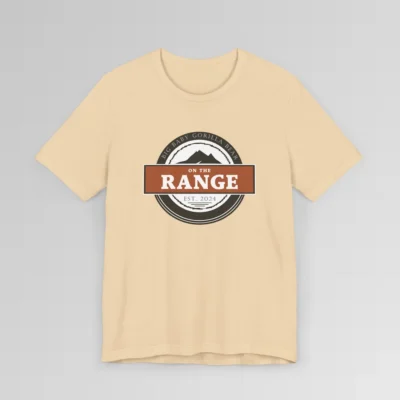 The front of a shirt with a dark brown, and cream white circular design, with the words "Big Baby Gorilla Bear" wrapped on the top part of the circle, and "Est. 2024" on the bottom inner part of the circle. In the center, there is an orange and brown rectangle with the words "On The Range", and a mountain graphic. Color = Soft Cream.