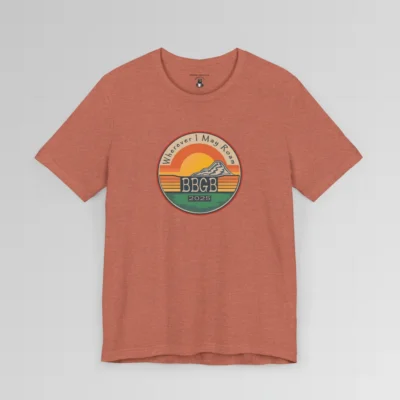 The front of a heather clay tee. with a circular, retro sunset design, with mountains, the letters "BBGB", "2025" in the middle, and the words "Wherever I May Roam" on the top of the circle. Red, Deep blue, teal, orange, yellow, and cream colors in the design.