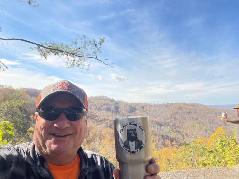 A male wearing a hat and sunglasses, with a tin mug that has the Big Baby Gorilla Bear Logo sticker, and rolling hills in the background on a sunny day.